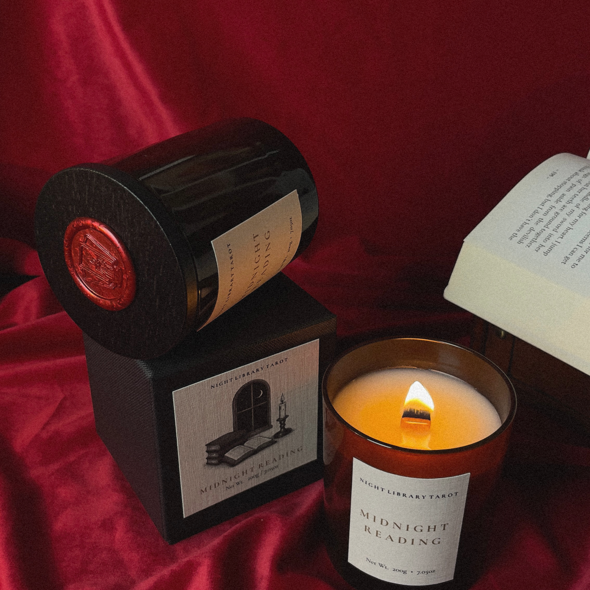 A candle product set on a rich red satin fabric background, featuring a black jar with a red wax seal resting on its black box, alongside a lit amber glass candle and an open book. The labels read 'Midnight Reading' and 'The Library Tarot'.
