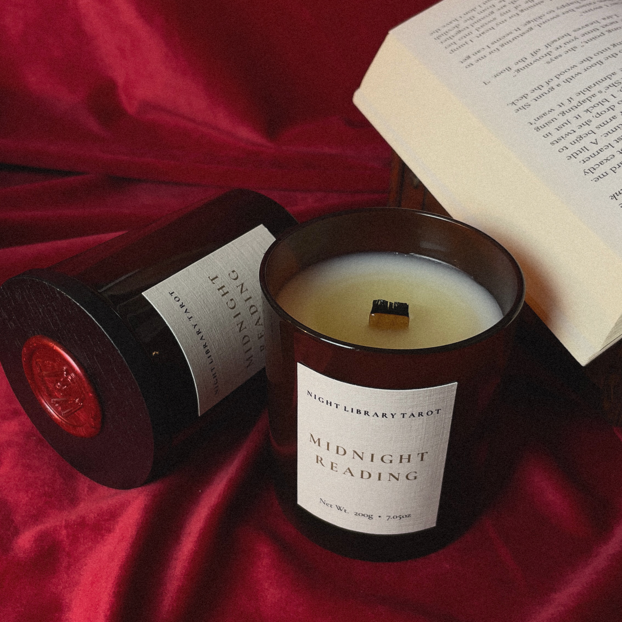 A dark amber glass candle jar labeled 'Midnight Reading' by Night Library Tarot, shown with its matching lid beside it on a deep red satin fabric, with an open book in the background.