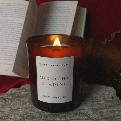 An amber glass jar candle with a lit flame sits on a stone surface in front of an open book, with a dark red fabric background and a brown leather-bound book visible behind it. The front label reads 'NIGHT LIBRARY TAROT' and 'MIDNIGHT READING'.