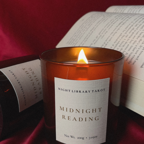 A lit amber glass candle labeled 'NIGHT LIBRARY TAROT MIDNIGHT READING' sits in front of an open book on a dark red fabric background.