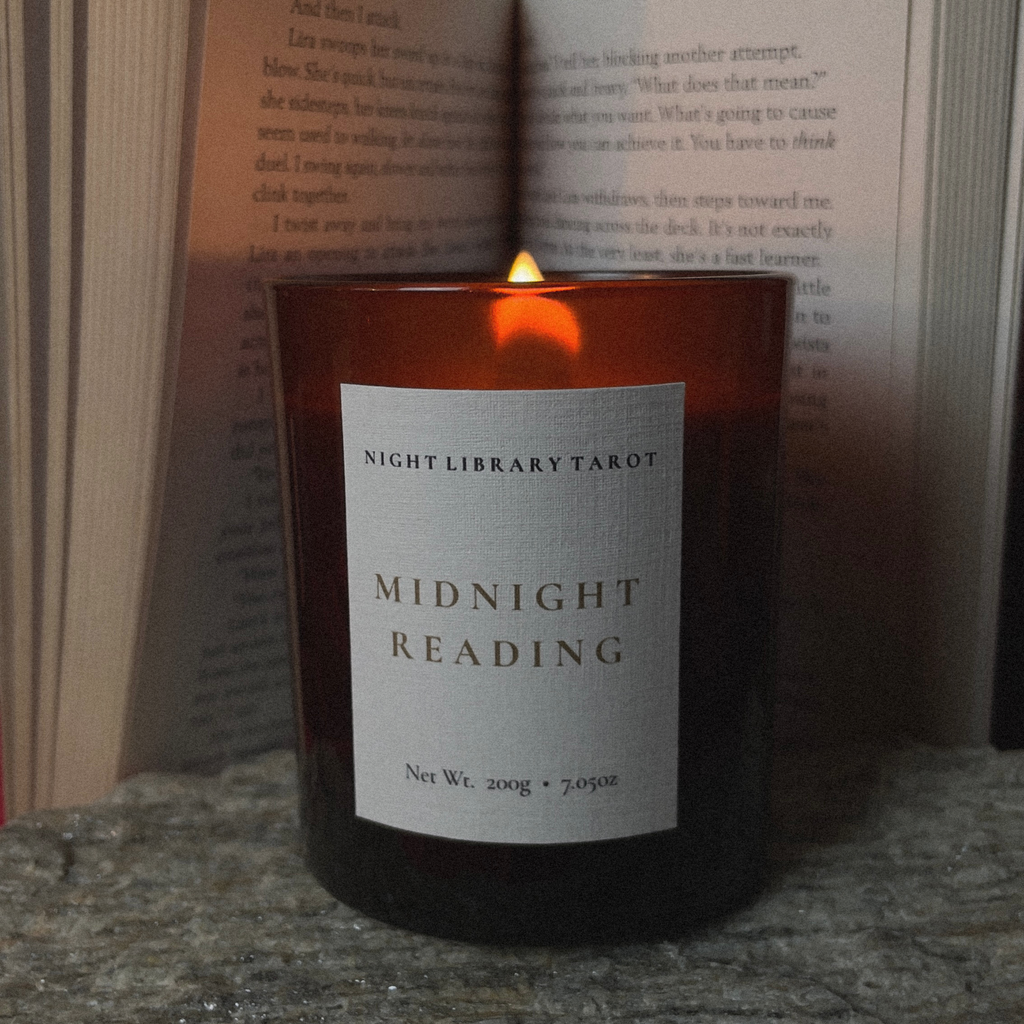 A lit amber glass candle with a white label reading 'NIGHT LIBRARY TAROT MIDNIGHT READING' sits on a stone surface in front of open books.