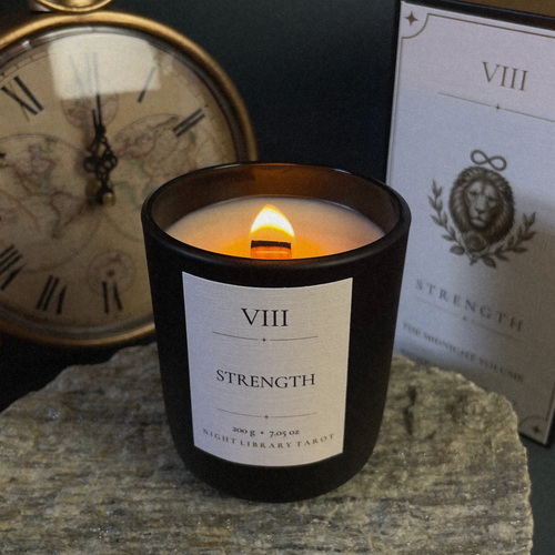 A black glass candle jar with a white label reading 'VIII STRENGTH' sits lit on a stone surface, with a vintage clock and a printed card in the background.