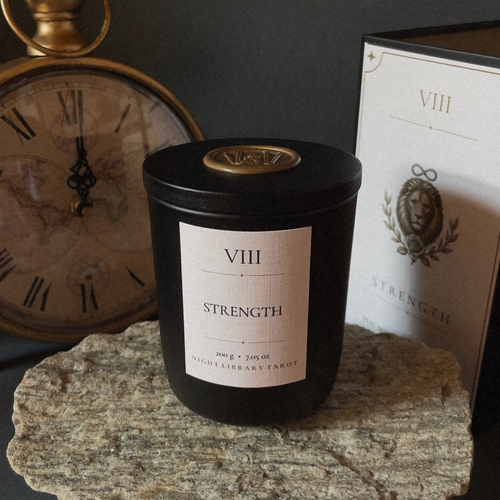 A black candle jar labeled 'VIII STRENGTH' sits on a stone slab, with its branded box behind it and a vintage clock in the background.