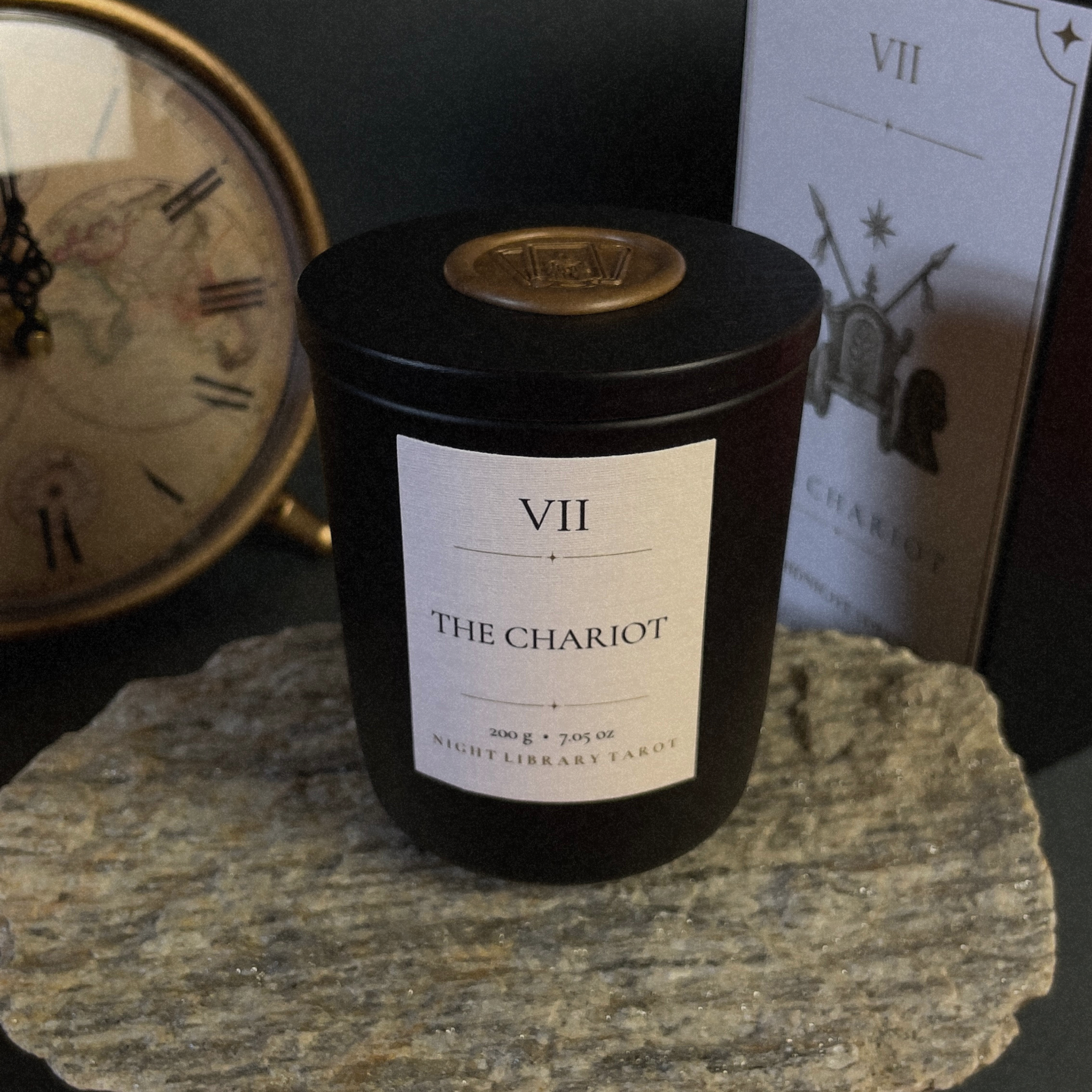 A black glass candle jar labeled 'VII THE CHARIOT' sits on a stone slab, with a vintage clock and a tarot card in the background.