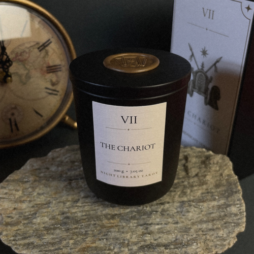 A black glass candle jar labeled 'VII THE CHARIOT' sits on a stone slab, with a vintage clock and a tarot card in the background.