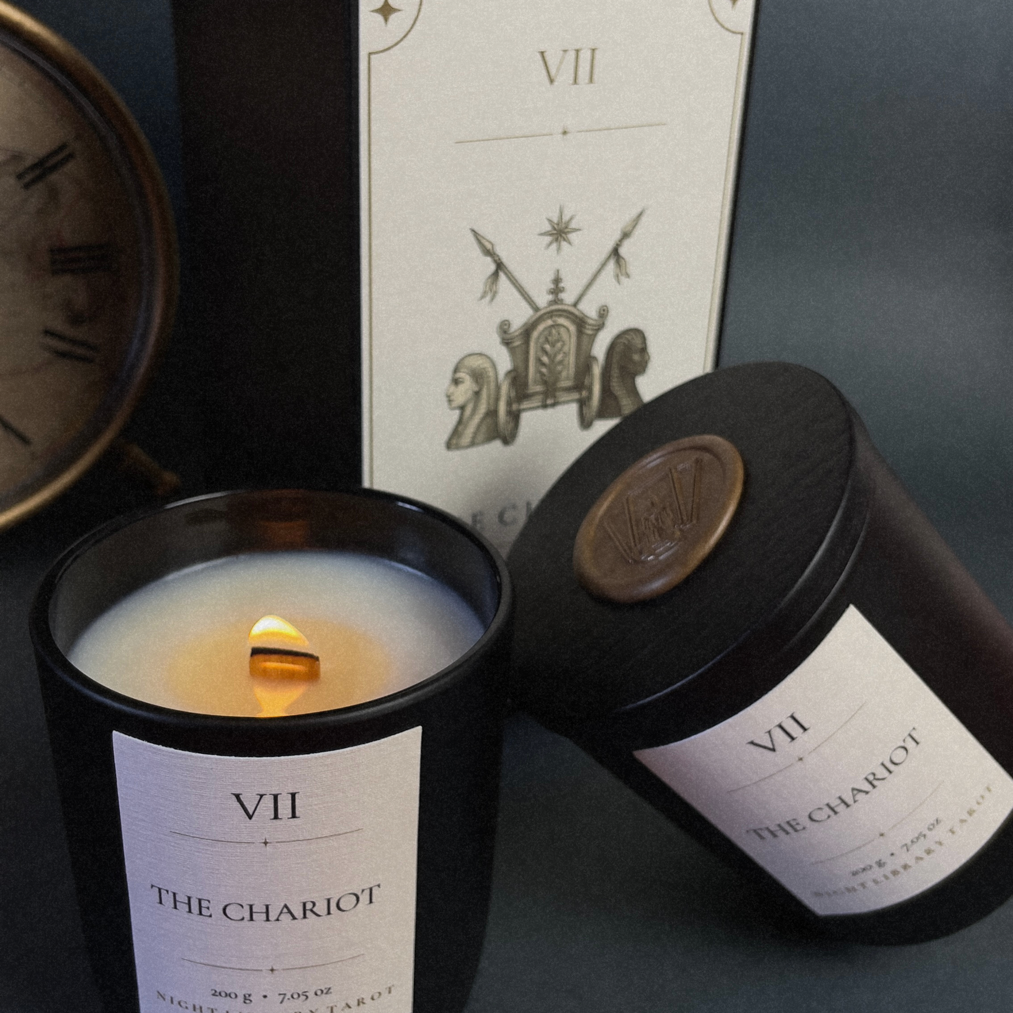 A black glass jar candle with a lit wick and white wax sits beside its matching black cylindrical tin and a tall product box labeled VII THE CHARIOT on a dark tabletop.