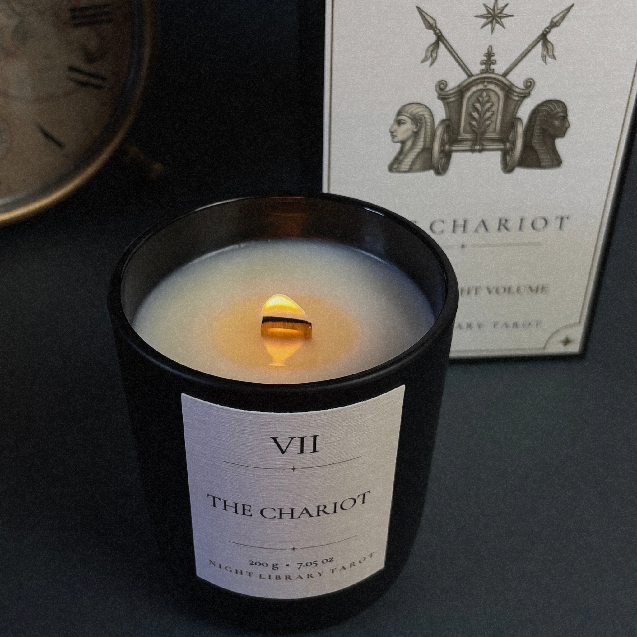 A lit black glass candle jar labeled 'VII THE CHARIOT' sits in front of its matching product box, which reads 'THE CHARIOT' with a chariot illustration. The scene is styled on a dark surface with a blurred clock in the background.