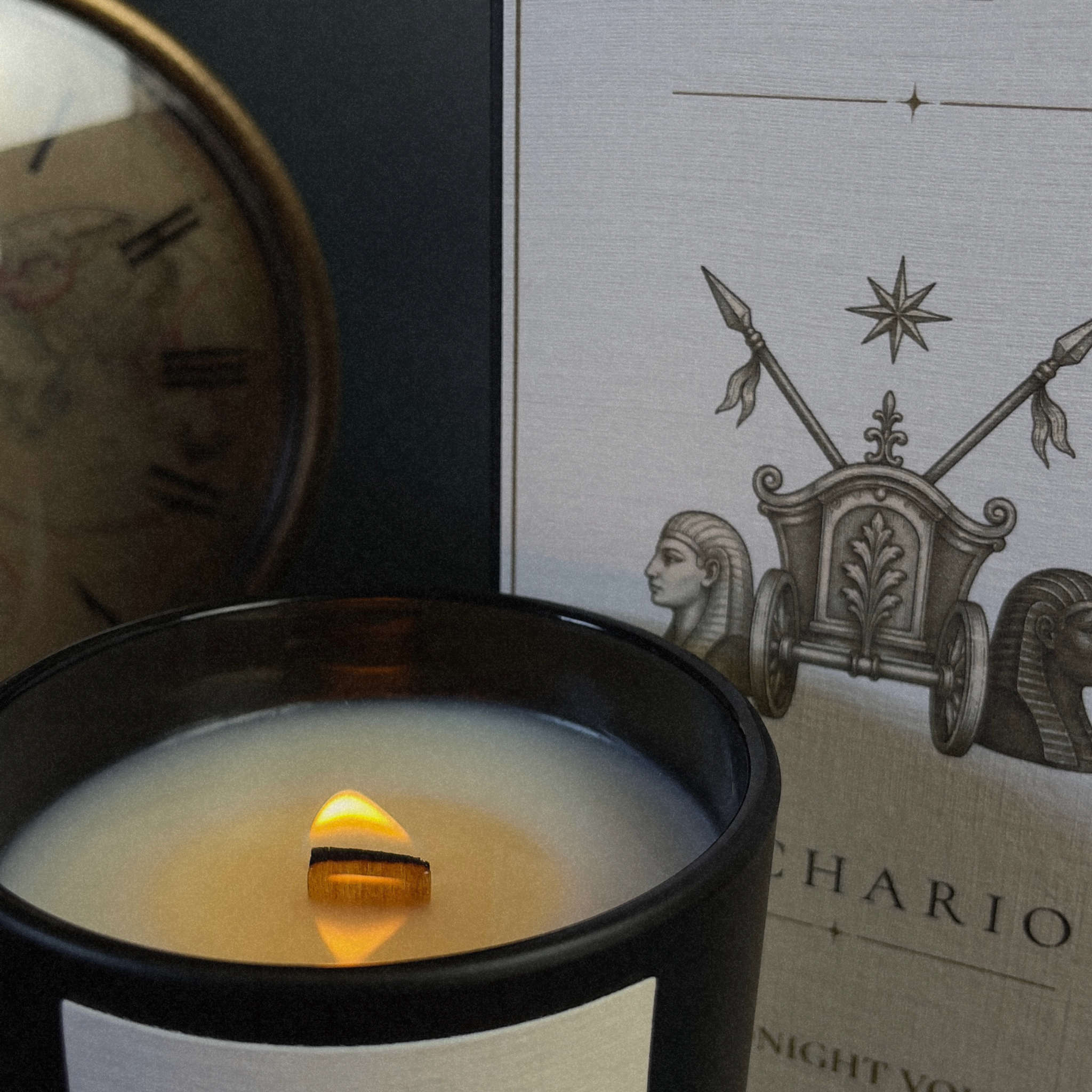 A close-up of a black glass candle jar with a single wooden wick lit and glowing, set against a dark background with part of a clock and a decorative printed card visible.