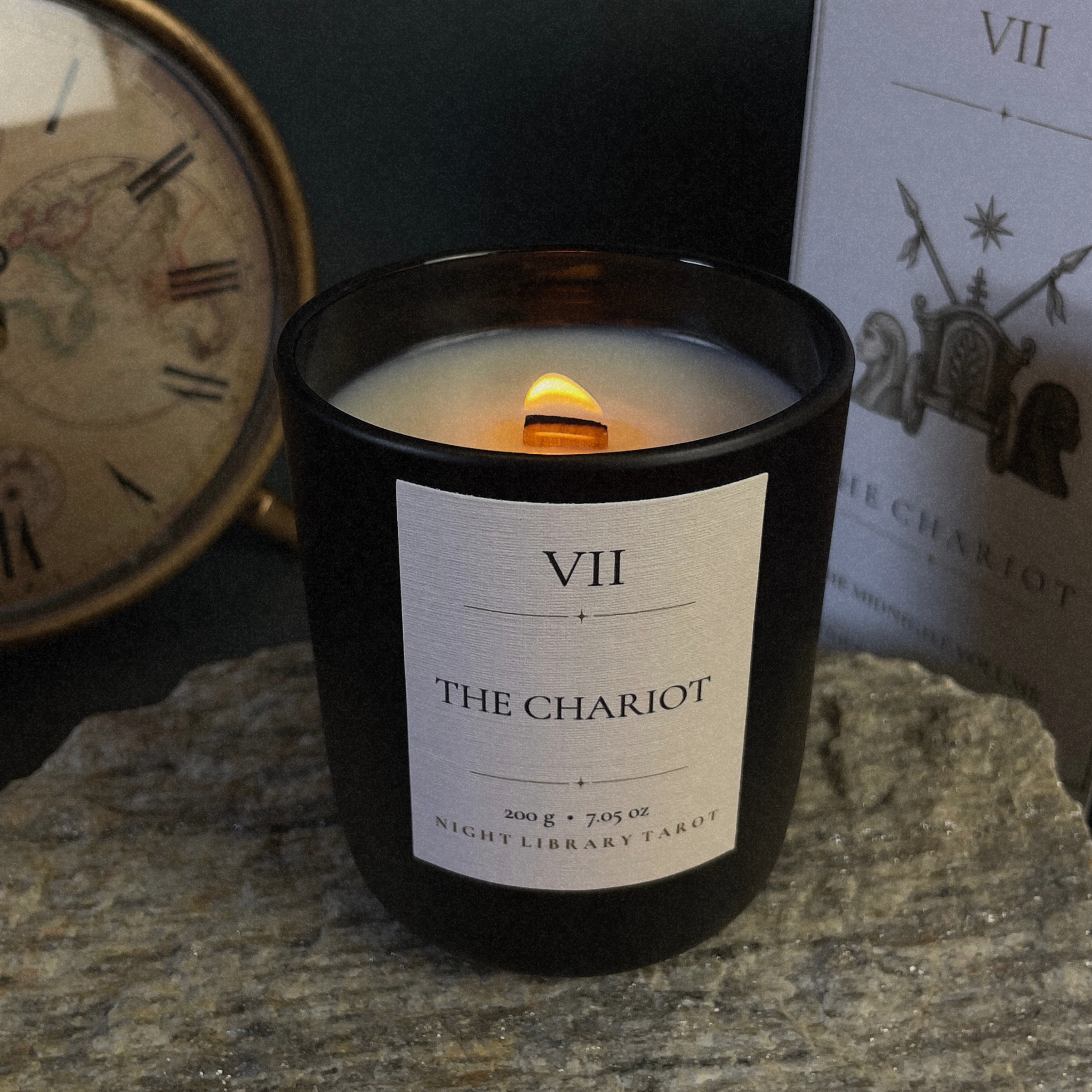 A black glass jar candle with a white label reading 'VII THE CHARIOT' sits lit on a stone surface, with a vintage clock and a printed card in the background.