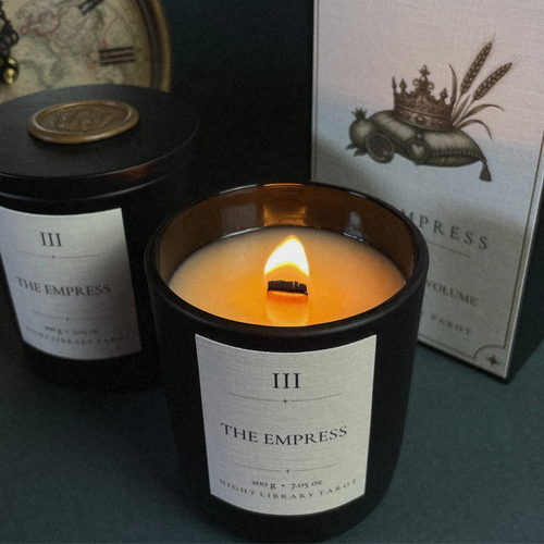 A lit candle in a black jar labeled 'THE EMPRESS' sits in front of its matching box and another unlit candle on a dark surface.
