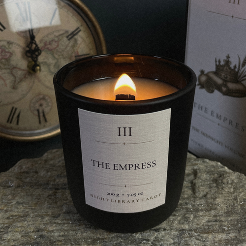 A lit candle in a matte black glass jar with a white label reading 'III THE EMPRESS NIGHT LIBRARY TAROT', placed on a stone surface with a vintage clock and book in the background.