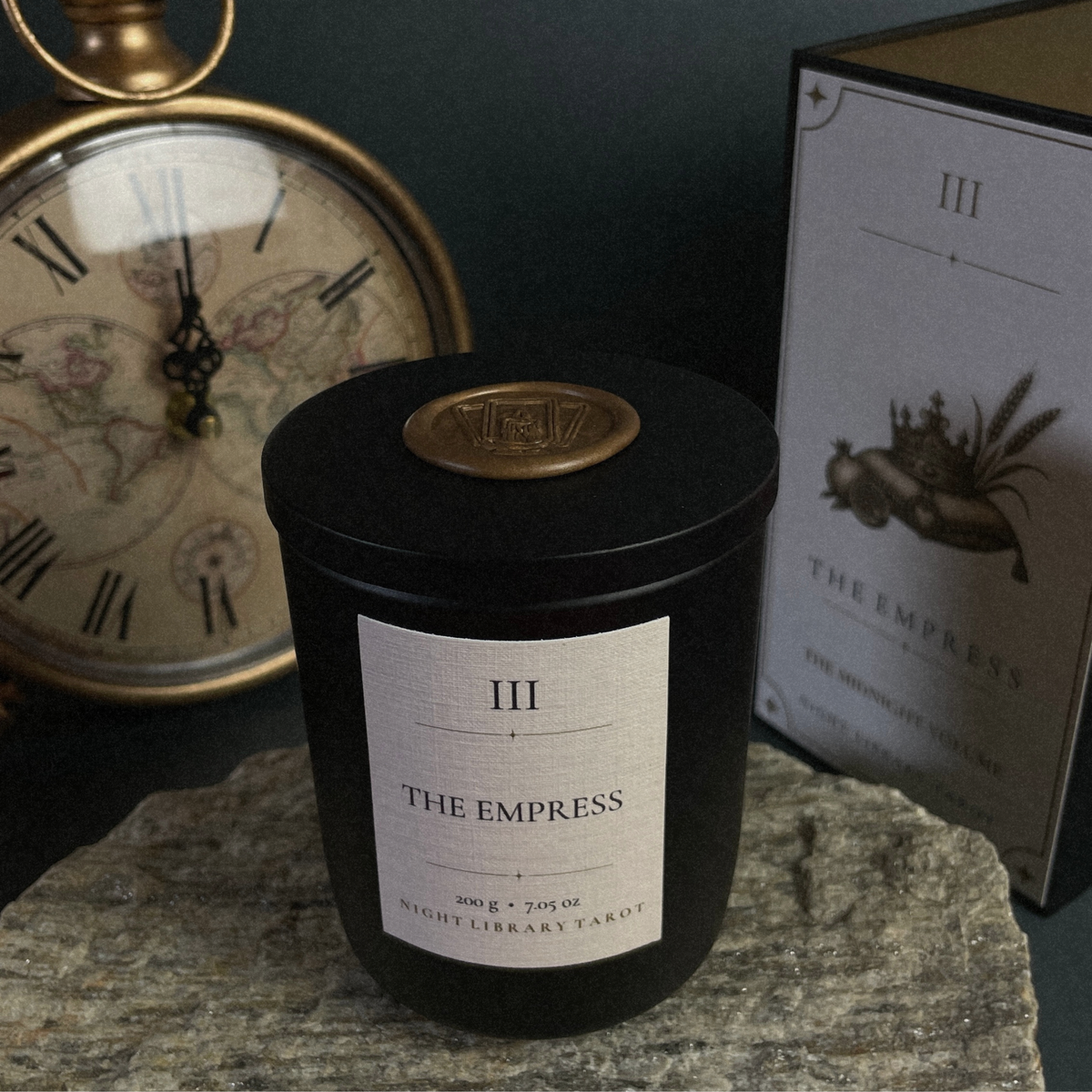 A black candle jar labeled 'III THE EMPRESS NIGHT LIBRARY TARIOT' sits on a stone surface, with a vintage brass clock and a book box in the background.