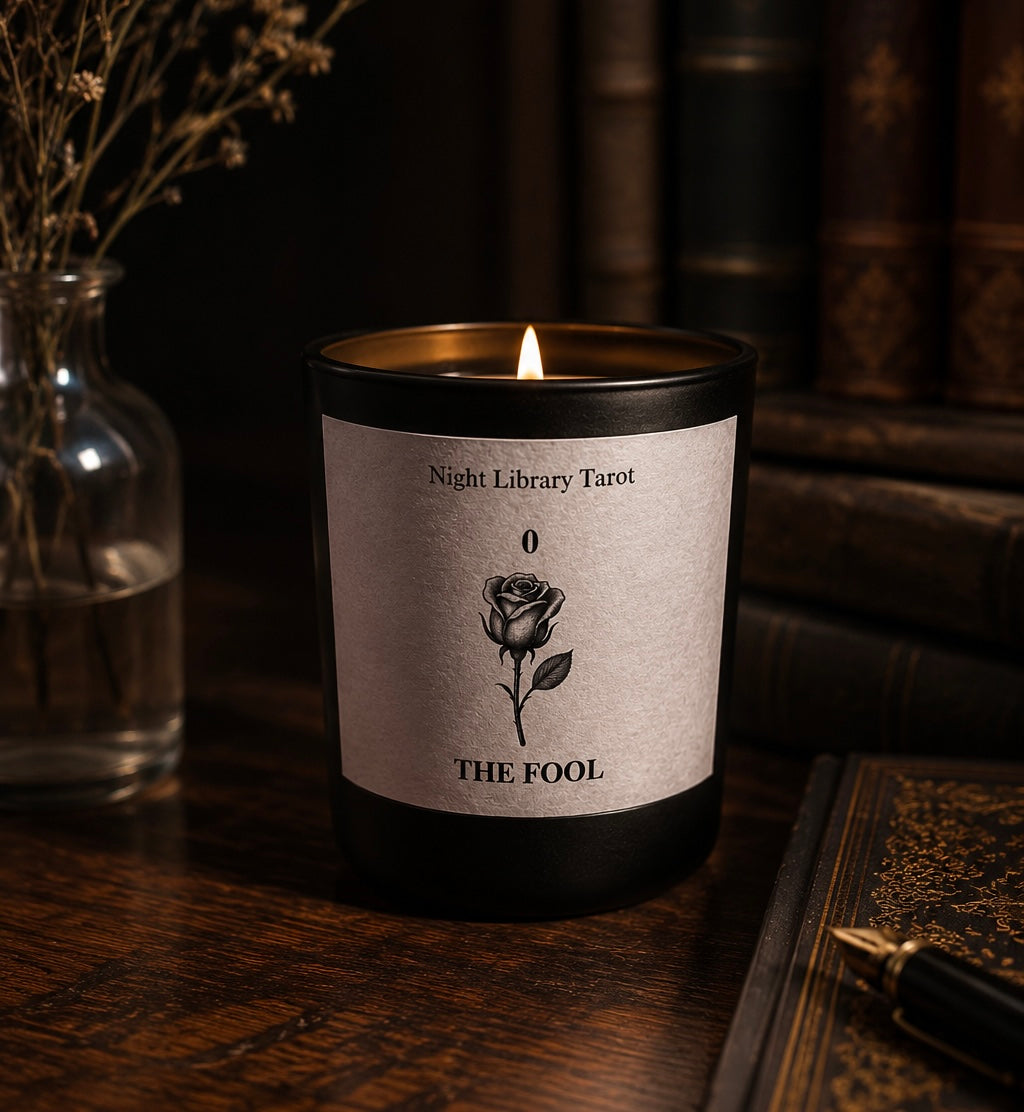 Candle with a rose design and 'The Fool' label on a dark surface, surrounded by books and a vase. The Fool tarot inspired luxury candle. Dark Academia.