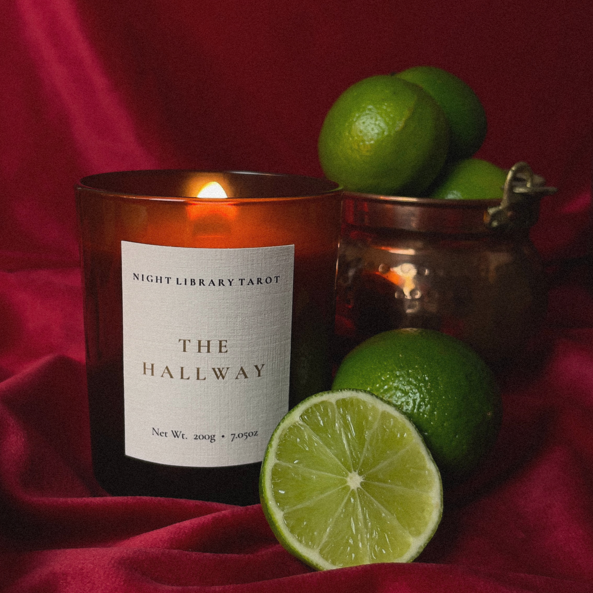 A lit amber glass candle labeled 'Night Library Tarot The Hallway' sits on draped red fabric beside whole and sliced limes, with a small copper-toned jar in the background.