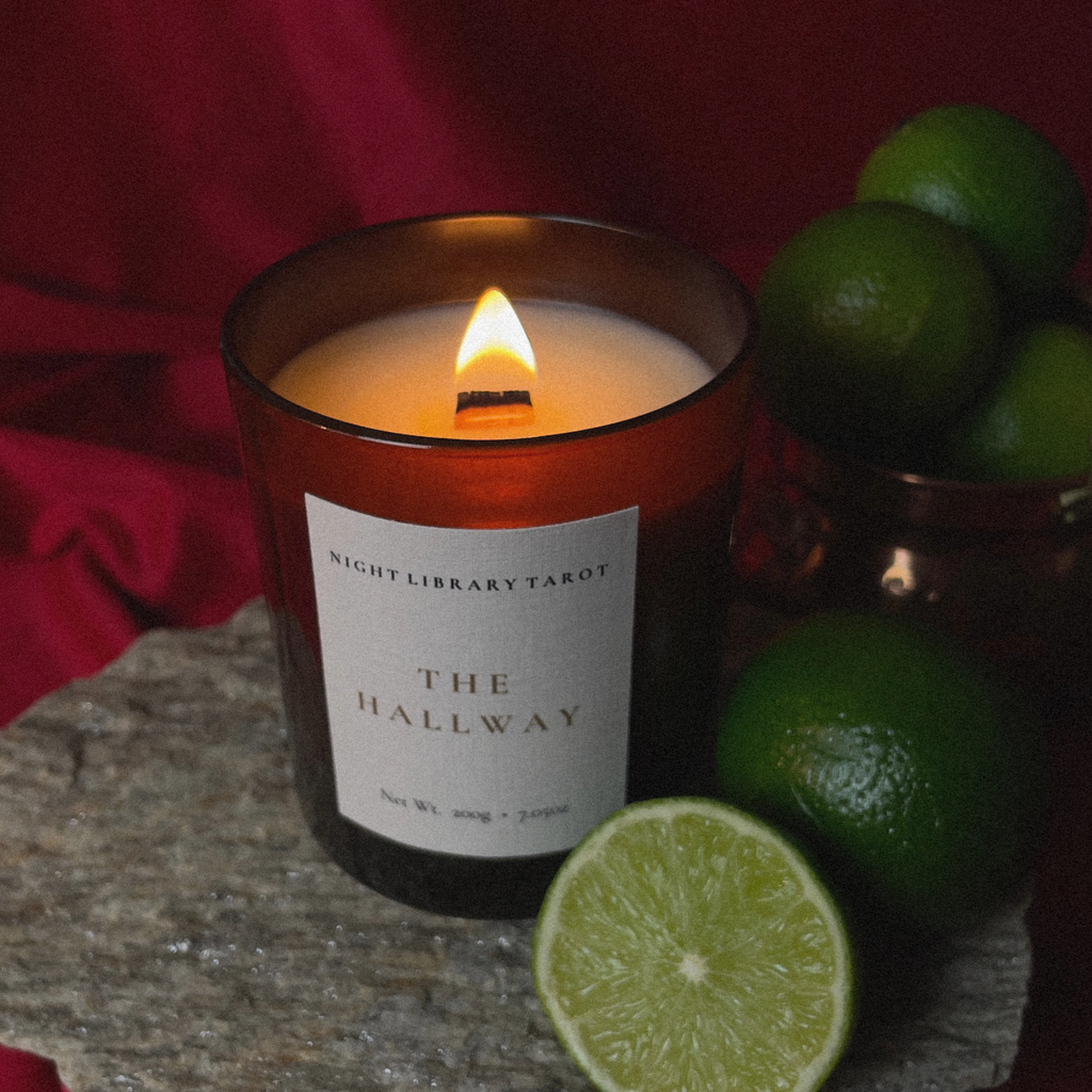 A lit amber glass candle with a white label reading 'NIGHT LIBRARY TAROT THE HALLWAY' sits on a stone surface, surrounded by limes and a red fabric backdrop.