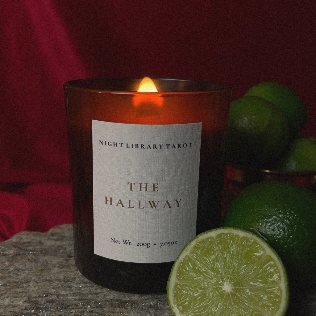 A lit amber glass candle labeled 'Night Library Tarot The Hallway' sits on a stone surface with limes in the foreground and a dark red draped background.