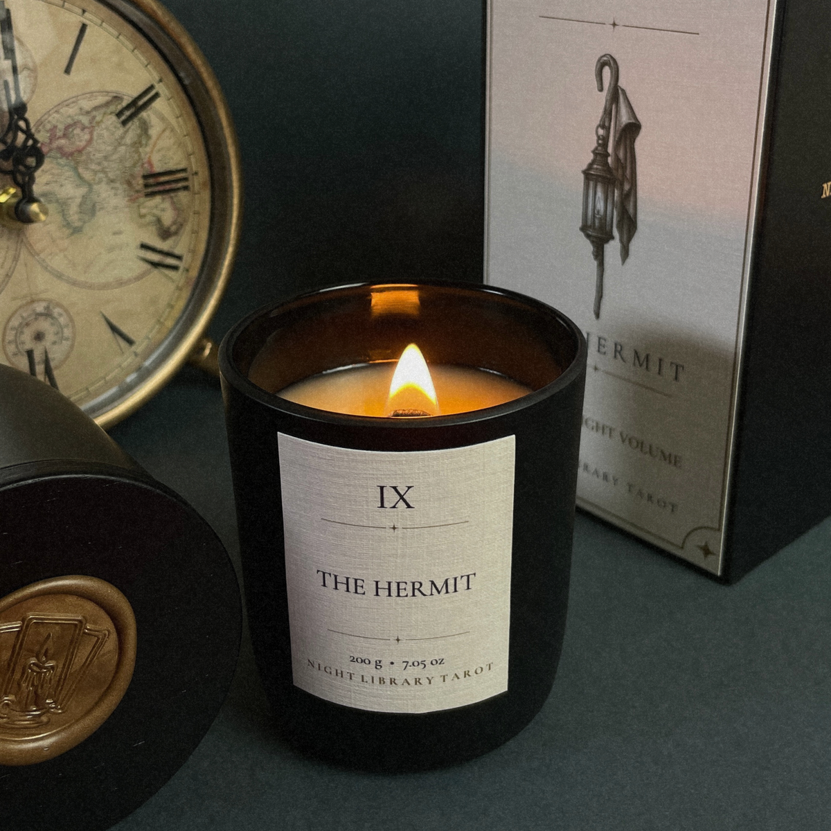 A lit black glass candle jar labeled 'IX THE HERMIT' sits on a dark tabletop with its matching box and a vintage clock in the background.