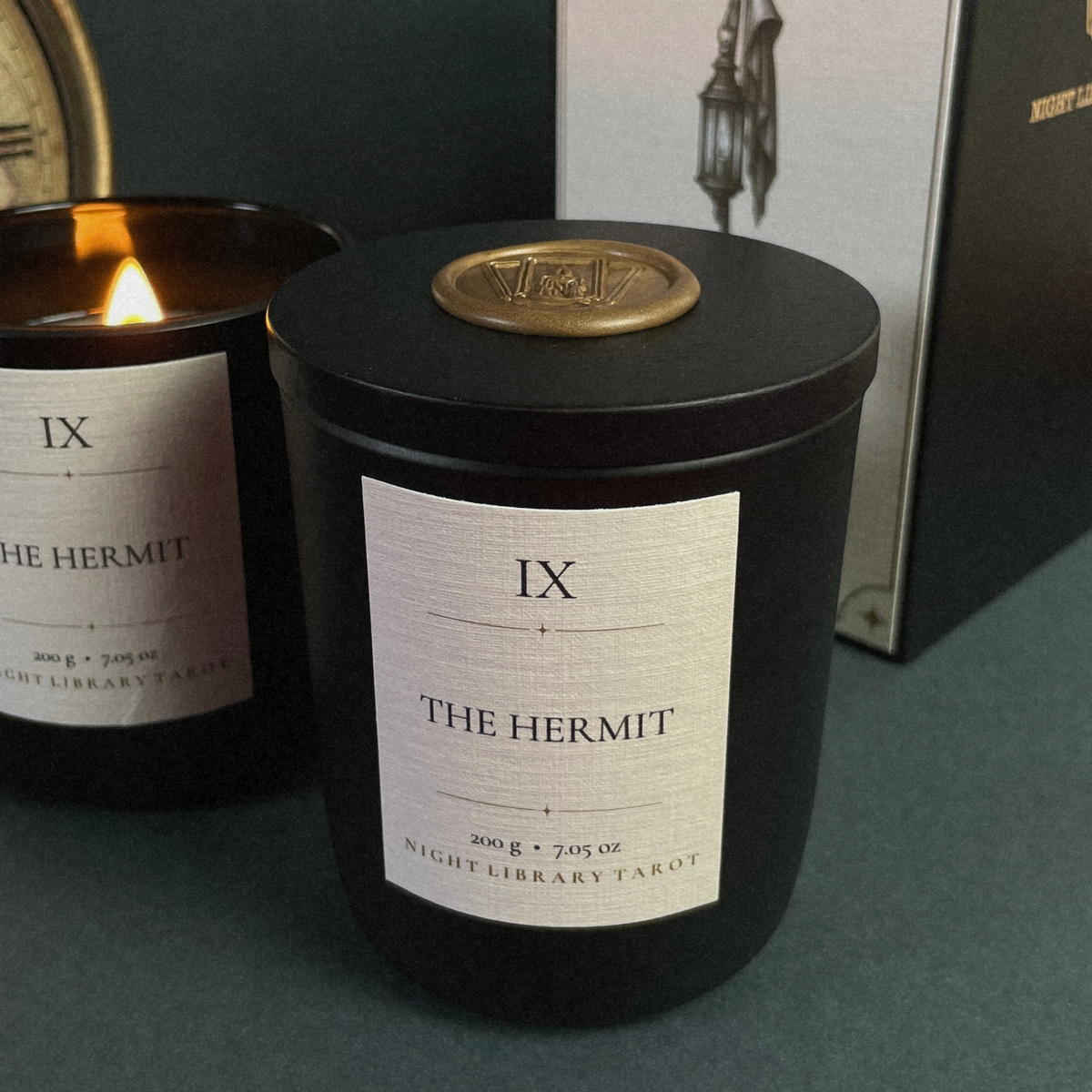 A black glass jar candle with a cream label reading 'IX THE HERMIT' and a gold seal on the lid, shown alongside another lit candle and a branded box in a moody setting.