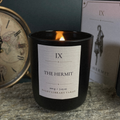A black glass candle jar with a lit flame and a white label reading 'IX THE HERMIT' and 'NIGHT LIBRARY TAROT', styled on a stone surface with a clock and a card box in the background.