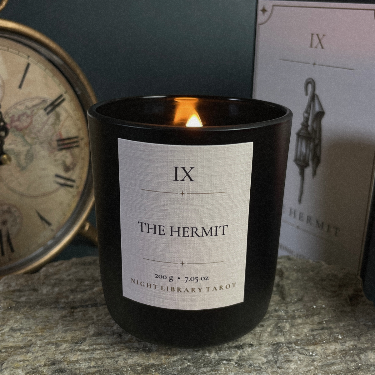 A black glass candle jar with a lit flame and a white label reading 'IX THE HERMIT' and 'NIGHT LIBRARY TAROT', styled on a stone surface with a clock and a card box in the background.