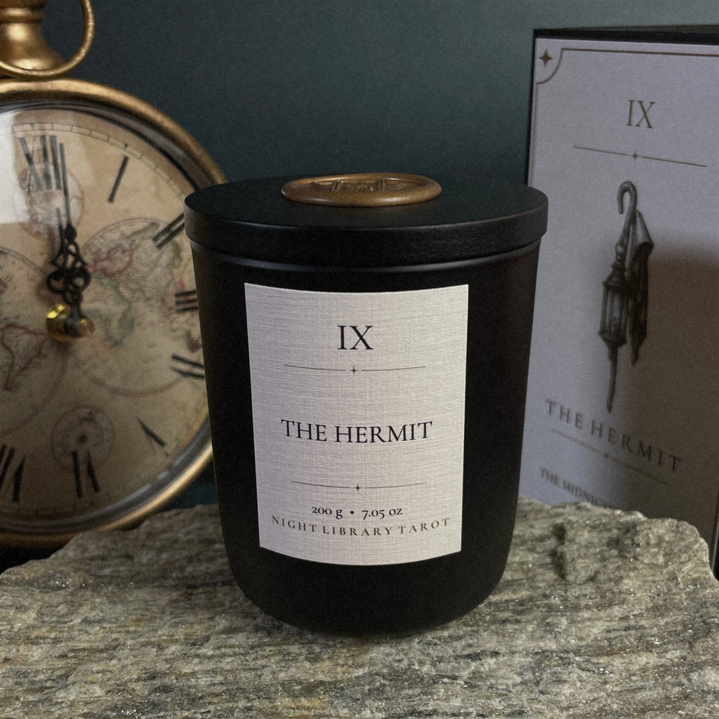 A black candle jar with a white label reading 'IX THE HERMIT' and 'NIGHTLIBRARY TAKOT' sits on a stone surface, with a vintage clock and a branded box in the background.