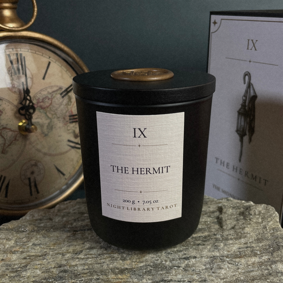 A black candle jar with a white label reading 'IX THE HERMIT' and 'NIGHTLIBRARY TAKOT' sits on a stone surface, with a vintage clock and a branded box in the background.
