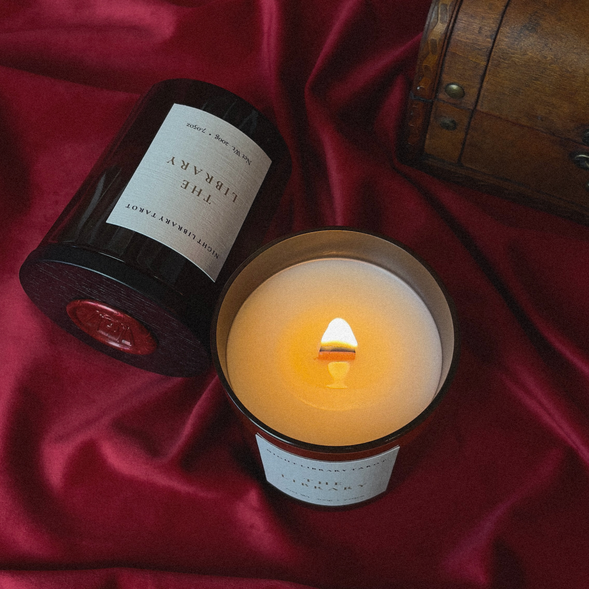 A lit candle in a clear glass jar sits on rich red satin fabric next to its dark brown wooden lid, which features a white label reading 'THE LIBRARY' and 'SILKSYANDS CANDLE CO.'.
