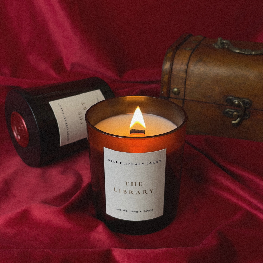 A lit amber glass candle jar labeled 'Night Library Tarot The Library' sits on draped red fabric, with a second candle lying nearby and a vintage wooden trunk in the background.