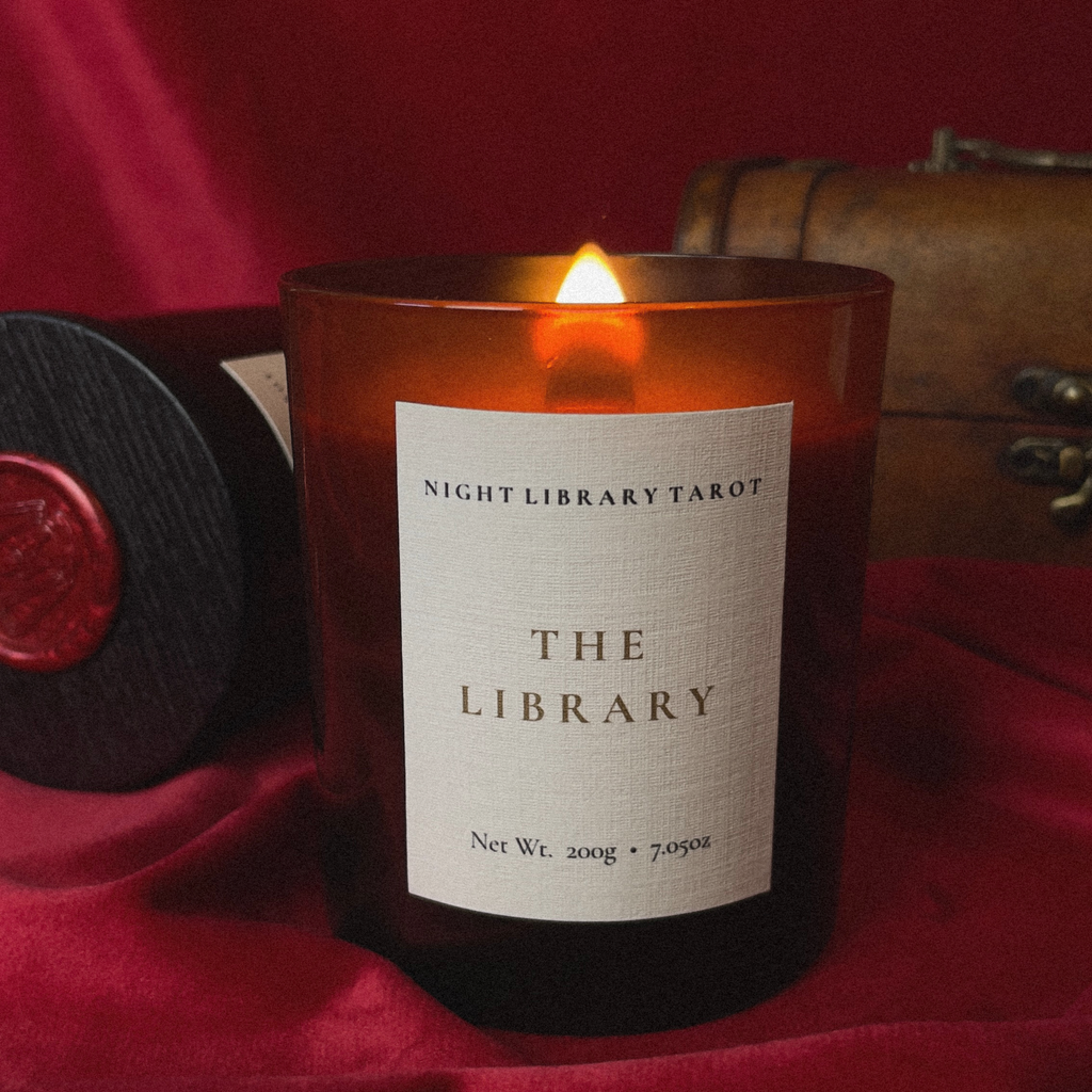 A lit amber glass candle jar labeled 'NIGHT LIBRARY TAROT THE LIBRARY' sits on draped red fabric, with a dark round box and a vintage-style leather case blurred in the background.