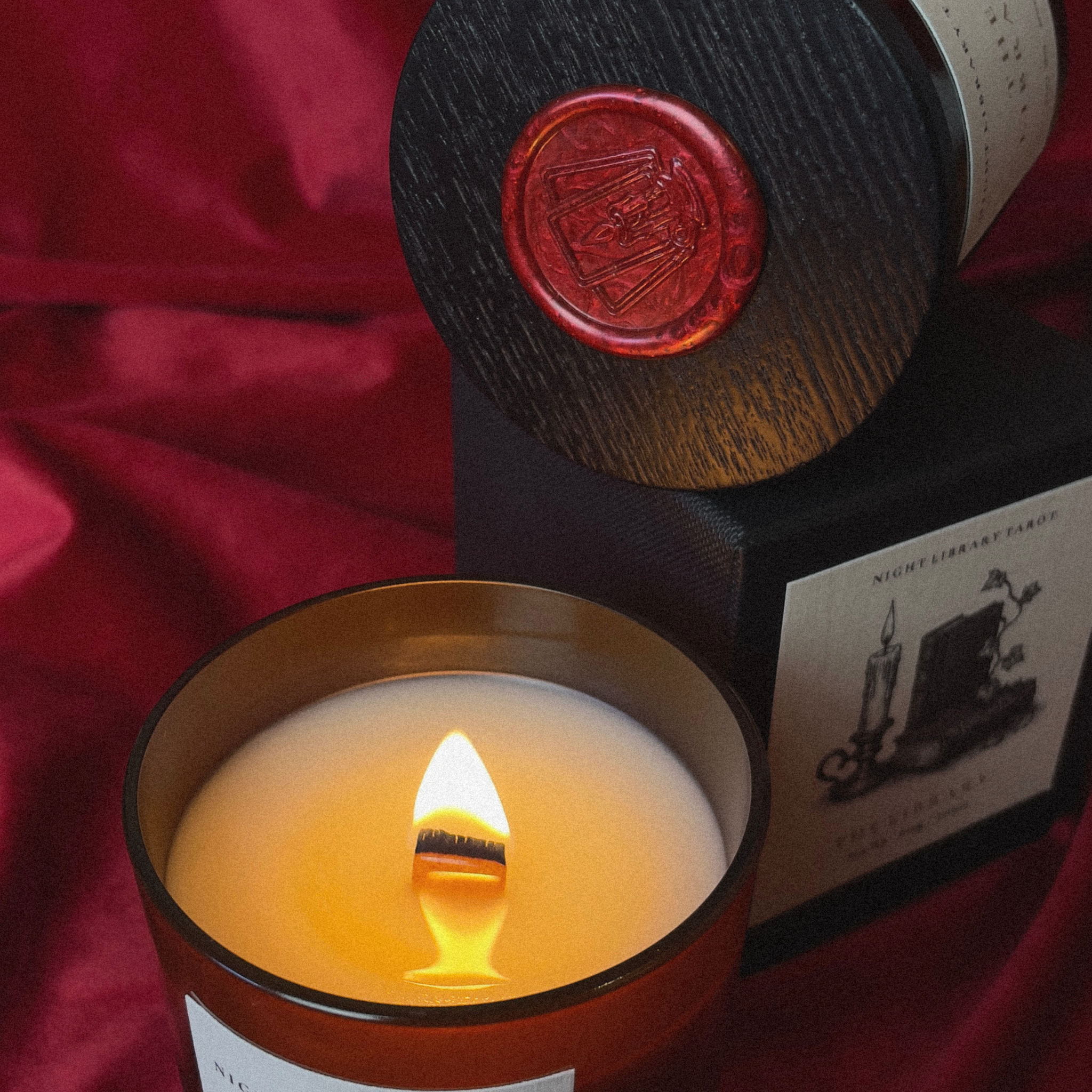 A lit candle in a dark amber glass jar with a single wick, shown in front of its black box packaging on draped red fabric. The lid leans against the box, featuring a red wax seal.