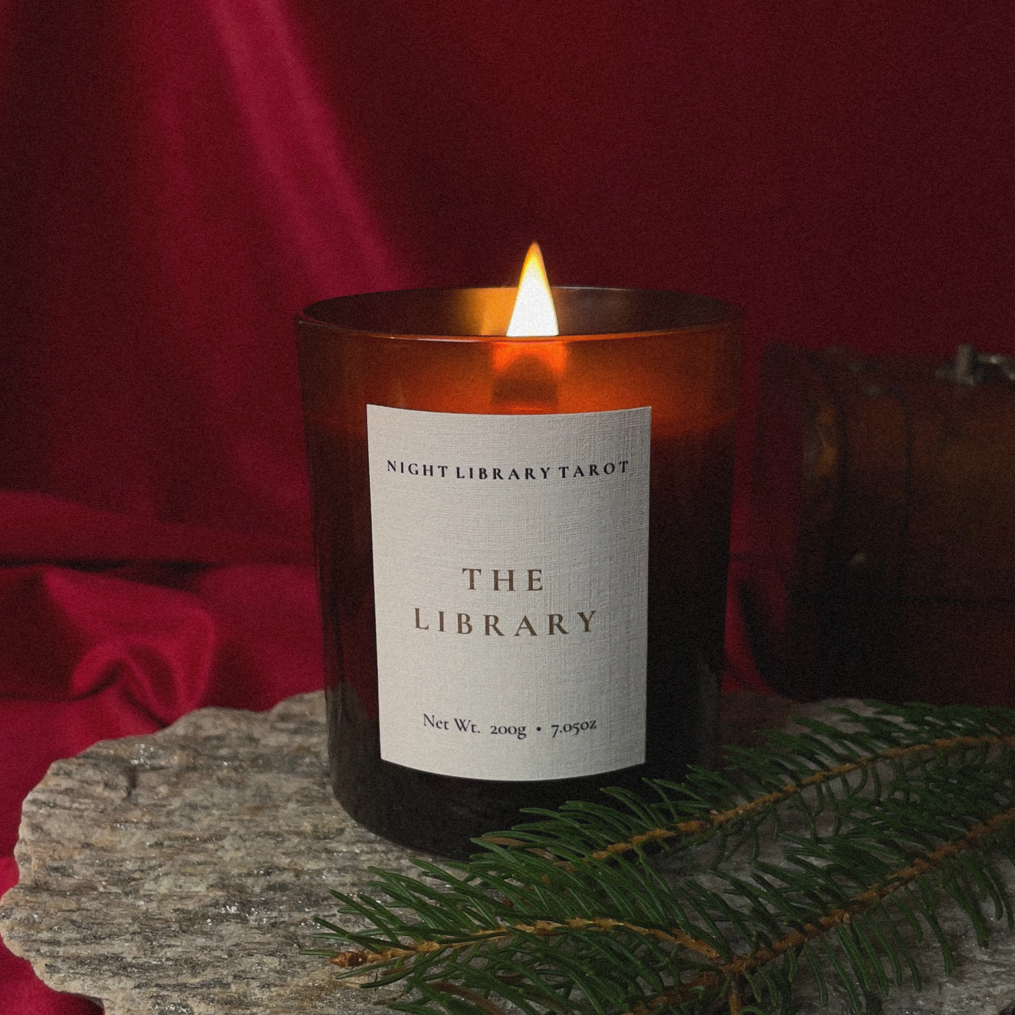 A lit amber glass candle jar with a white label reading 'NIGHT LIBRARY TAROT THE LIBRARY' sits on a stone slab with red fabric in the background and pine branches in the foreground.