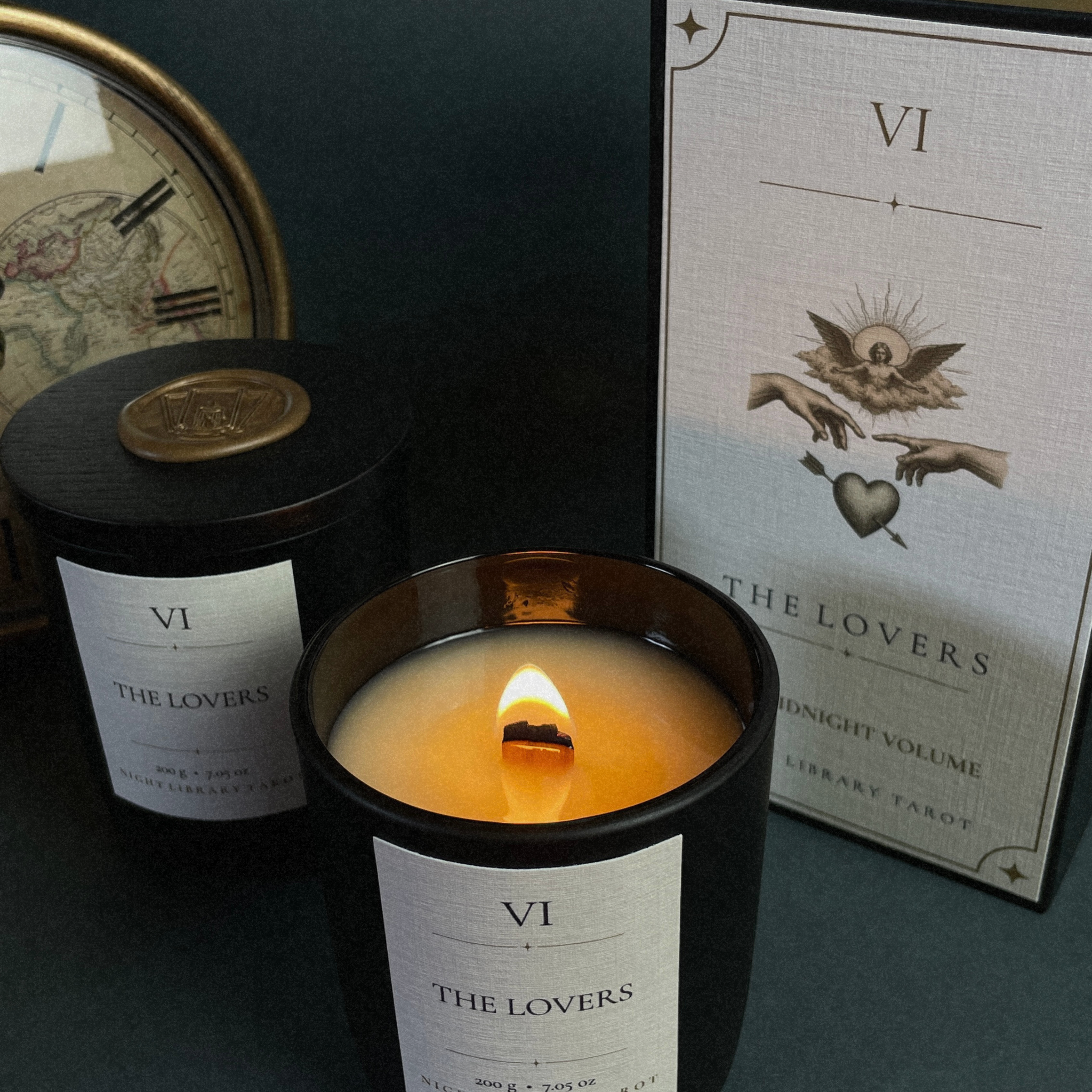 A dark candle jar with a lit flame in the foreground, alongside its matching box and a second unlit candle. The packaging features a white label with Roman numeral VI and the title 'THE LOVERS', with a library card design.