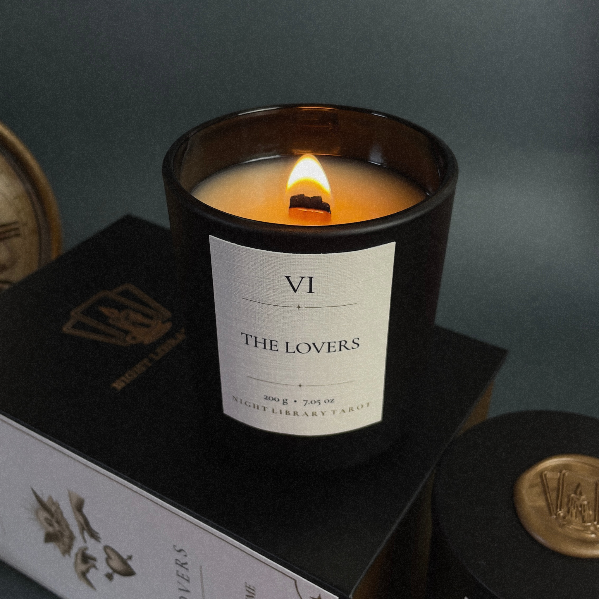 A lit candle in a dark amber glass jar sits on top of a black book or box. The front label reads "VI THE LOVERS" and shows a warm cream label.