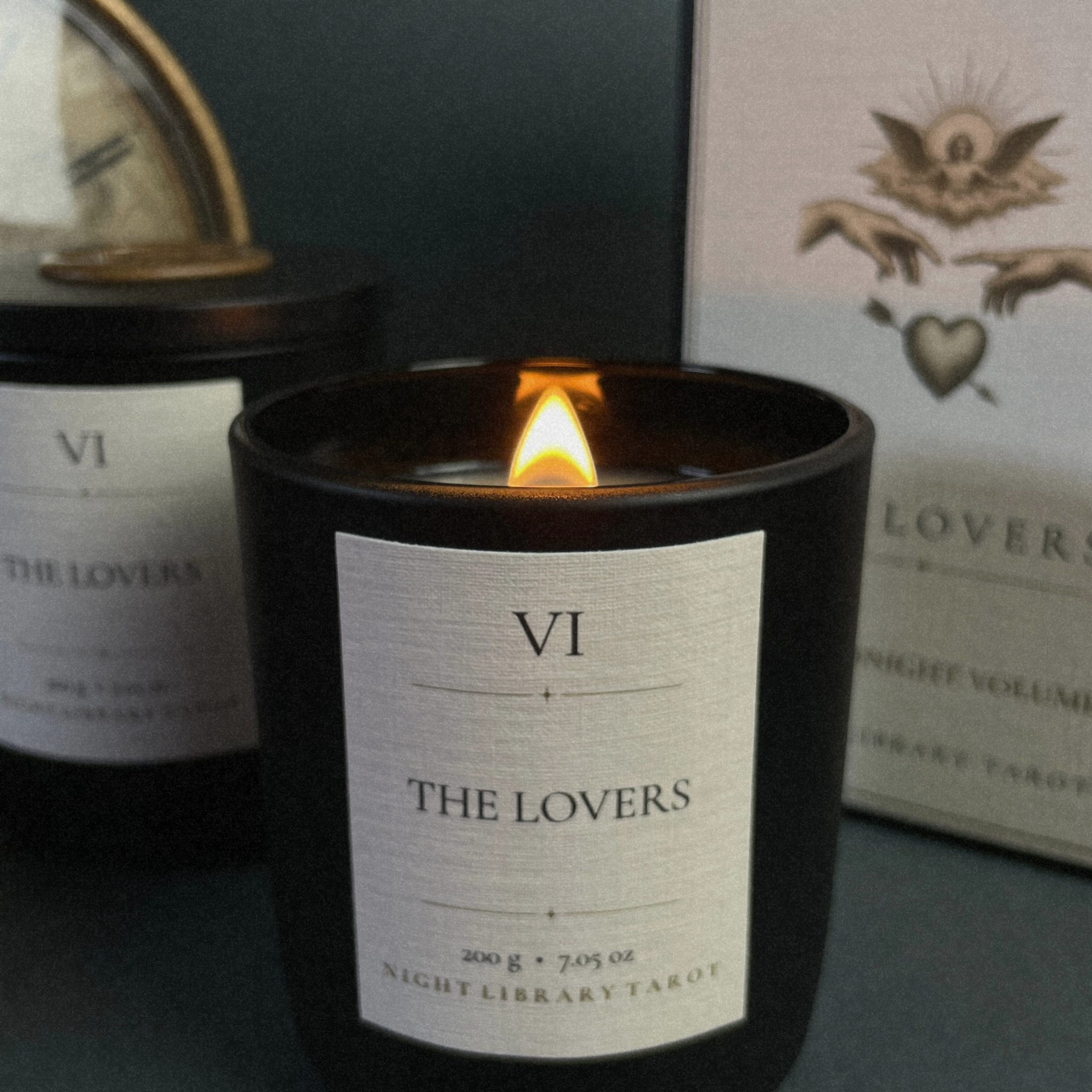 A black glass candle jar with a lit flame in the center, featuring a white label that reads 'VI THE LOVERS NIGHT LIBRARY TAROT.' Two matching candles and a decorative card are visible in the softly blurred background.