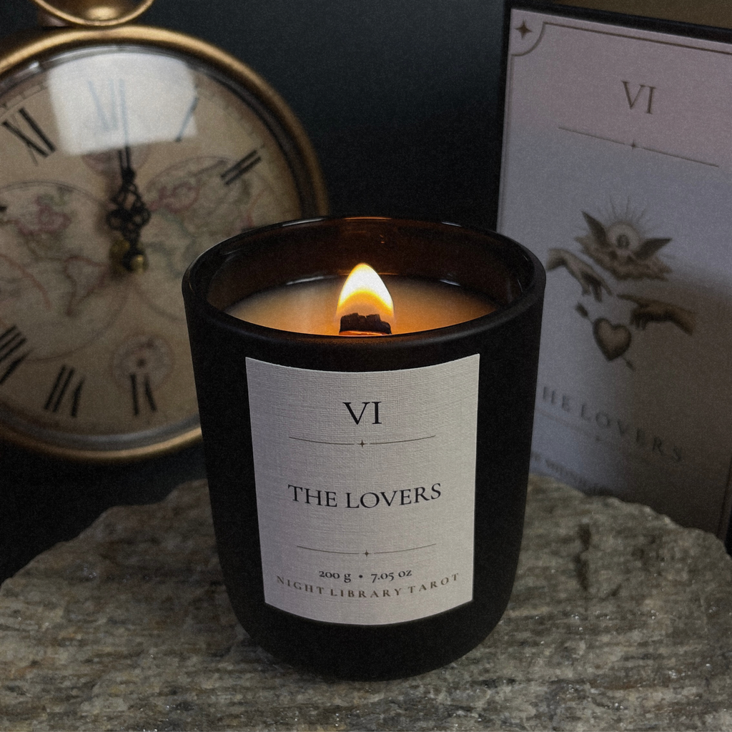 A black glass jar candle with a white label reading 'VI THE LOVERS' sits lit on a stone surface, with a vintage clock and a tarot card box in the background.