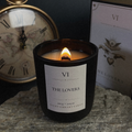 A black glass jar candle with a white label reading 'VI THE LOVERS' sits lit on a stone surface, with a vintage clock and a tarot card box in the background.
