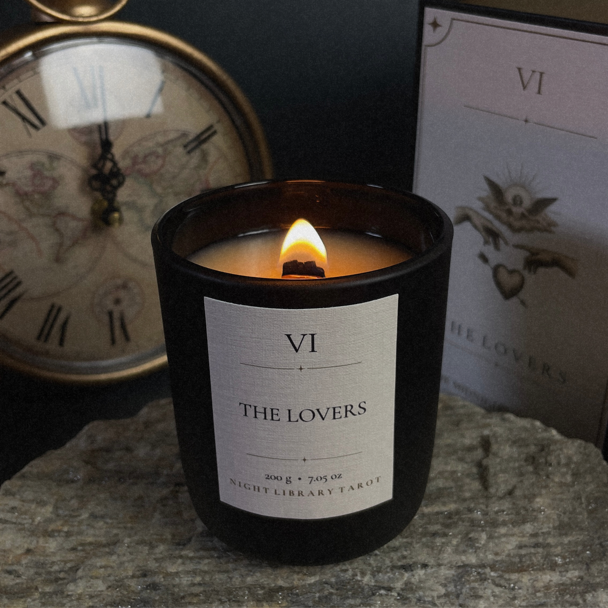 A black glass jar candle with a white label reading 'VI THE LOVERS' sits lit on a stone surface, with a vintage clock and a tarot card box in the background.