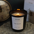 A black glass jar candle with a white label reading 'XVIII THE MOON NIGHT LIBRARY TAROT' lit with a visible flame, set on a stone surface with a clock and a card box in the background.