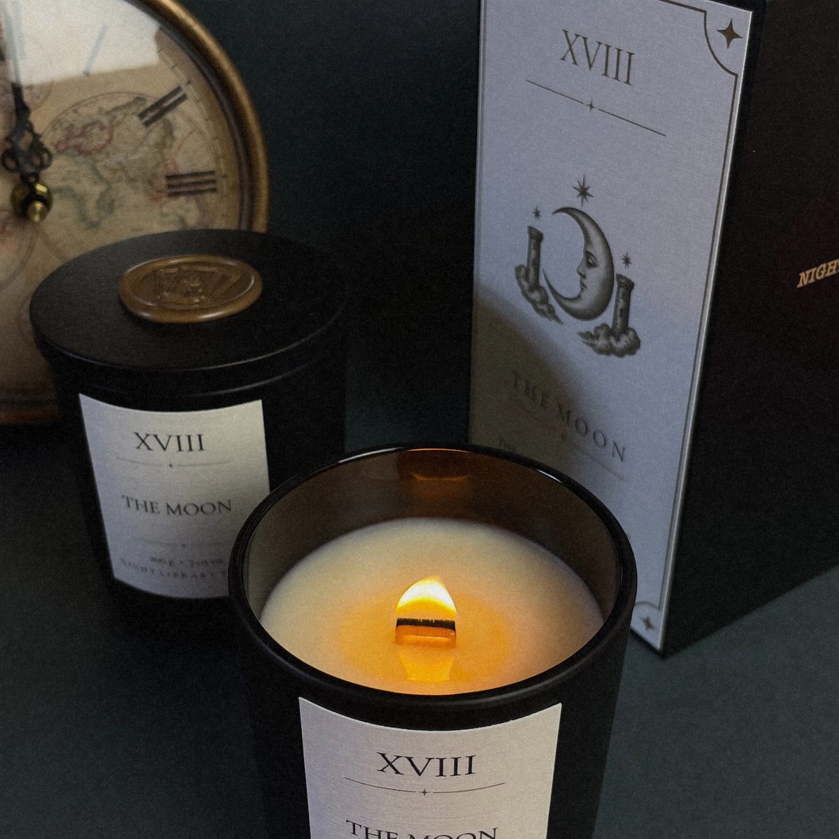 A black glass jar candle with a lit wick and cream wax is shown in the foreground, with a second unlit black candle and its box behind it. The packaging features a white label with a crescent moon and stars design and the text 'XVIII THE MOON'.
