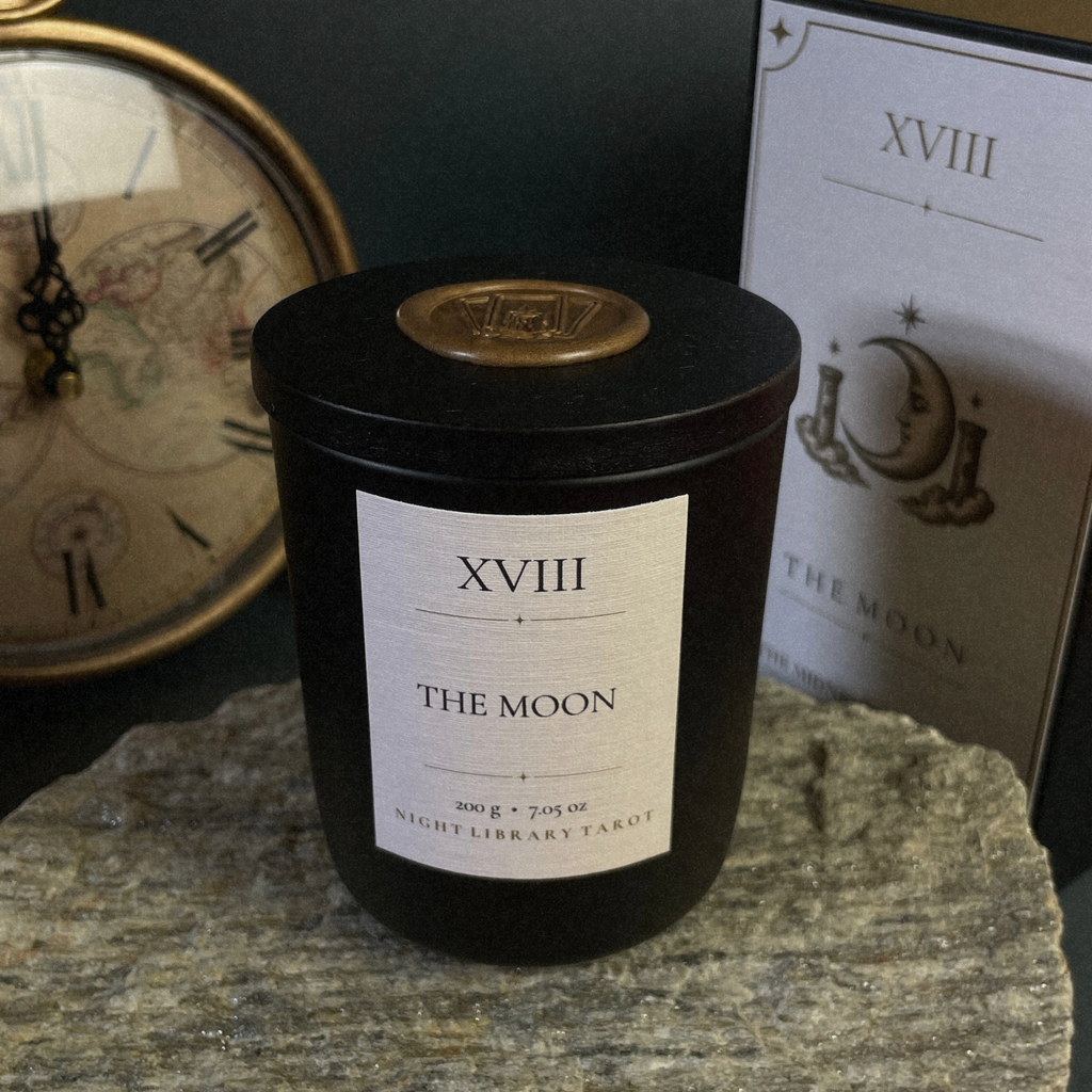 A black cylindrical candle jar labeled 'XVIII THE MOON' sits on a stone surface, with a matching card and a vintage-style clock in the background.