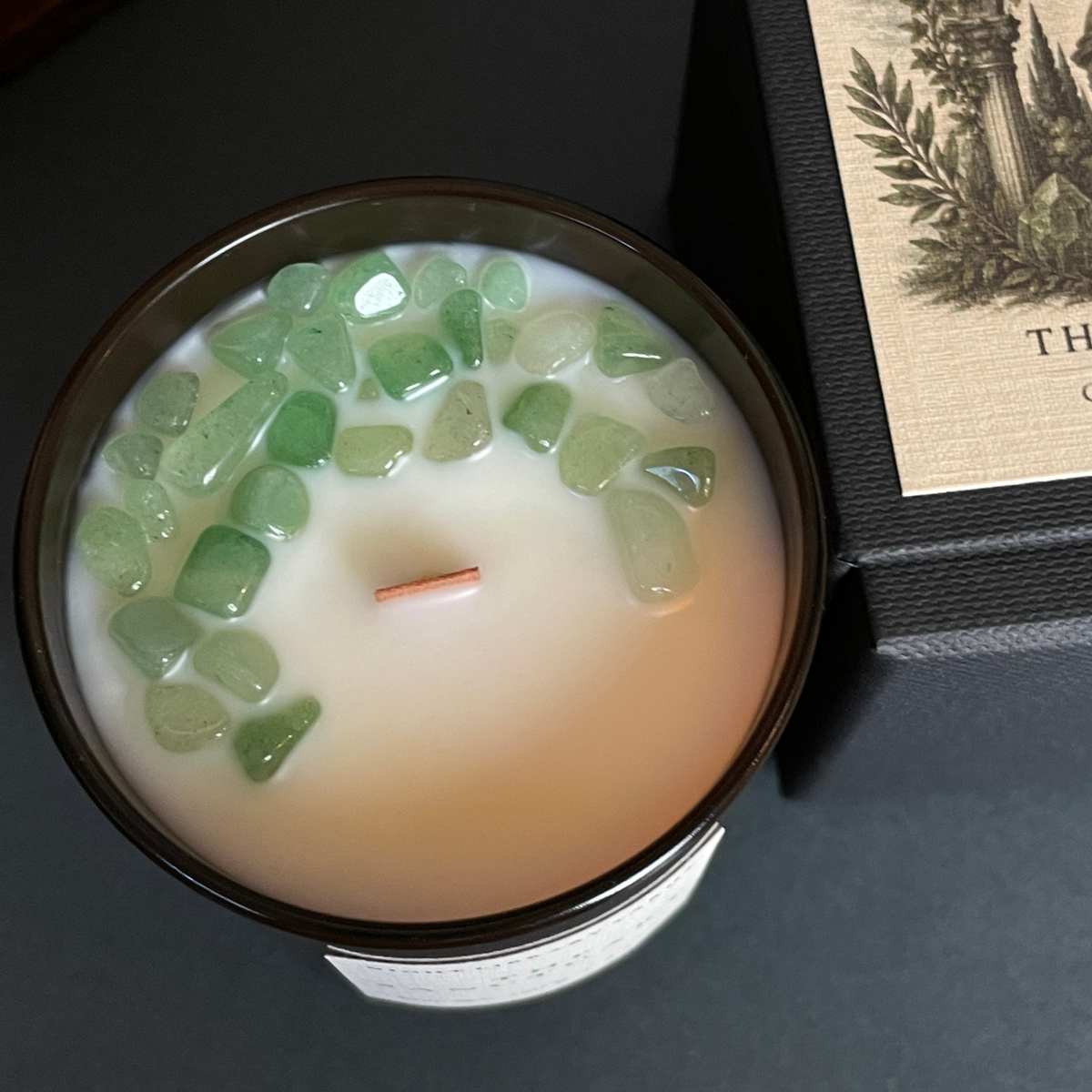 A top-down view of a candle in a dark brown glass jar filled with creamy off-white wax and decorated with green translucent stone chips. A portion of the black gift box with a beige label is visible on the right side.