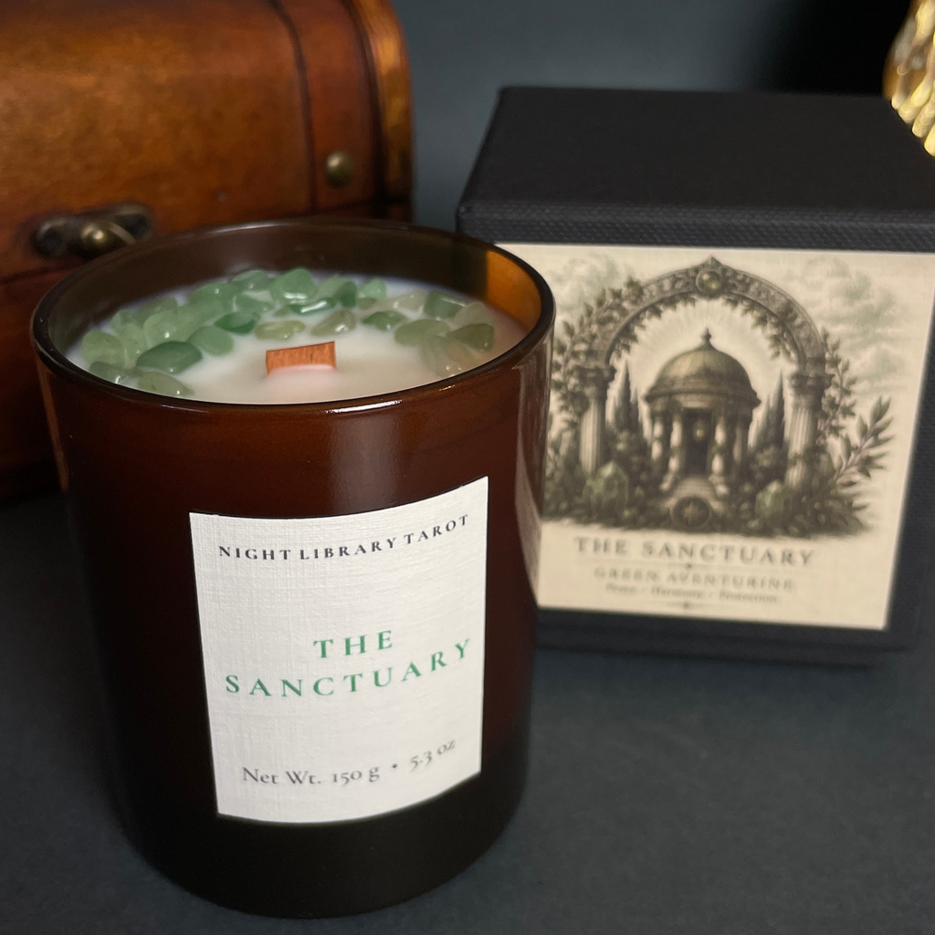 An amber glass jar candle labeled 'THE SANCTUARY' by Night Library Tarot, filled with white wax and topped with green stone chips, shown next to its black branded box on a dark surface.