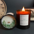 A lit amber glass candle labeled 'THE SANCTUARY' stands next to its open lid, which shows white wax and green stone accents. The candles are styled on a dark surface with a vintage clock and framed art in the background.