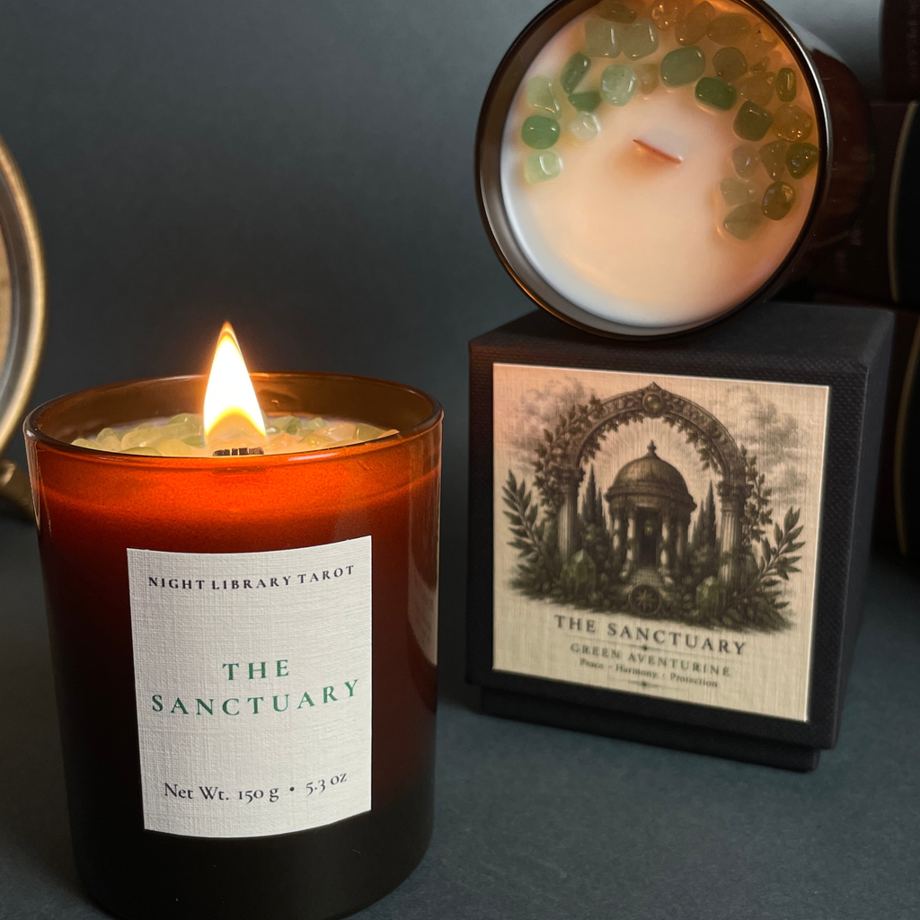 A lit amber glass candle labeled 'The Sanctuary' by Nightlibrary Tarot, shown beside its black box packaging and a second unlit candle with green stone accents on top.