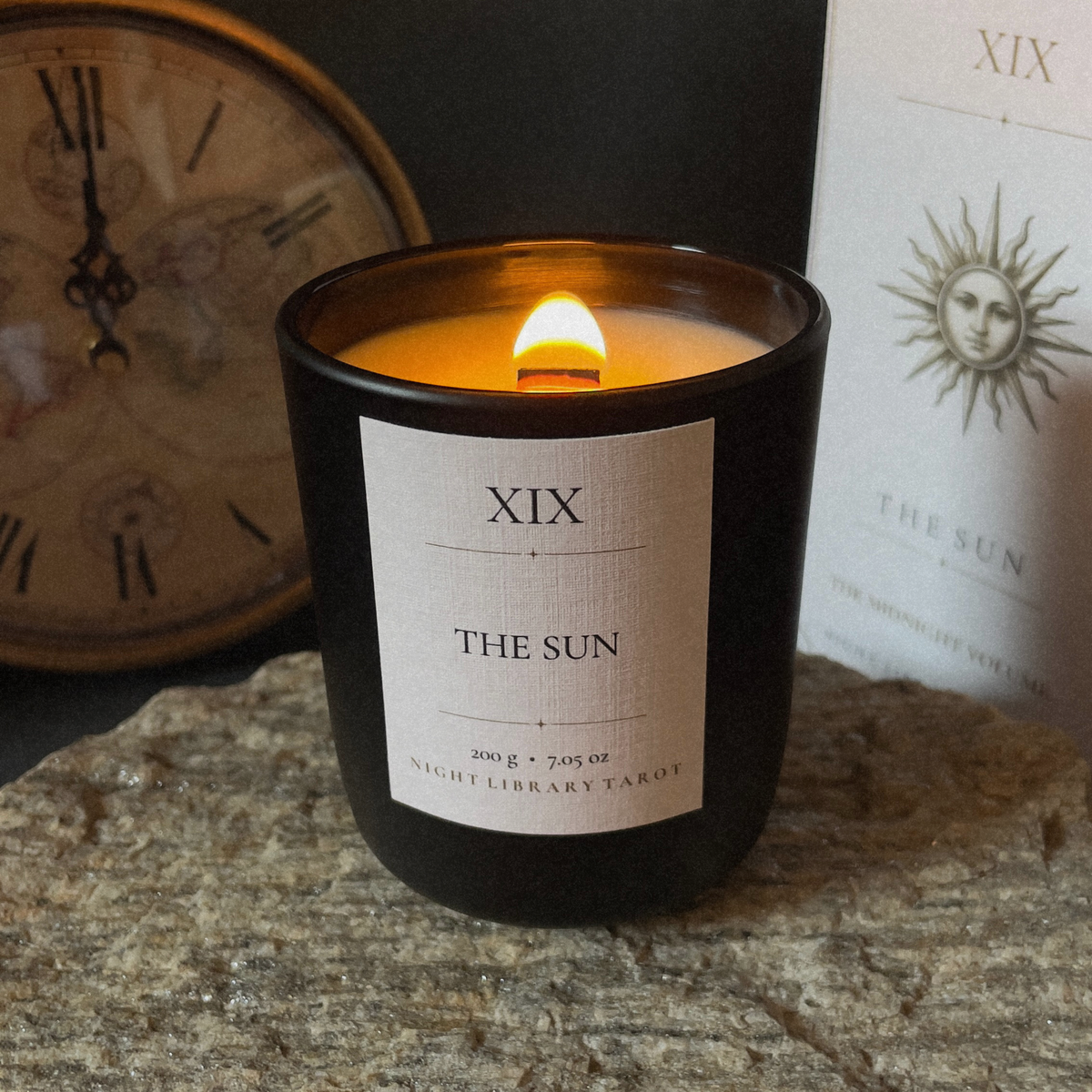 A lit black glass candle jar labeled 'XIX THE SUN' sits on a stone surface, with a vintage clock and a printed card in the background.