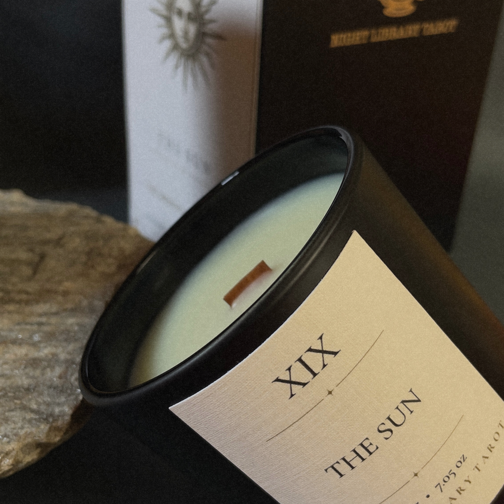 A close-up of a black candle jar labeled 'XIX THE SUN' with cream wax and a wooden wick, resting on a textured stone surface with a blurred dark box in the background.