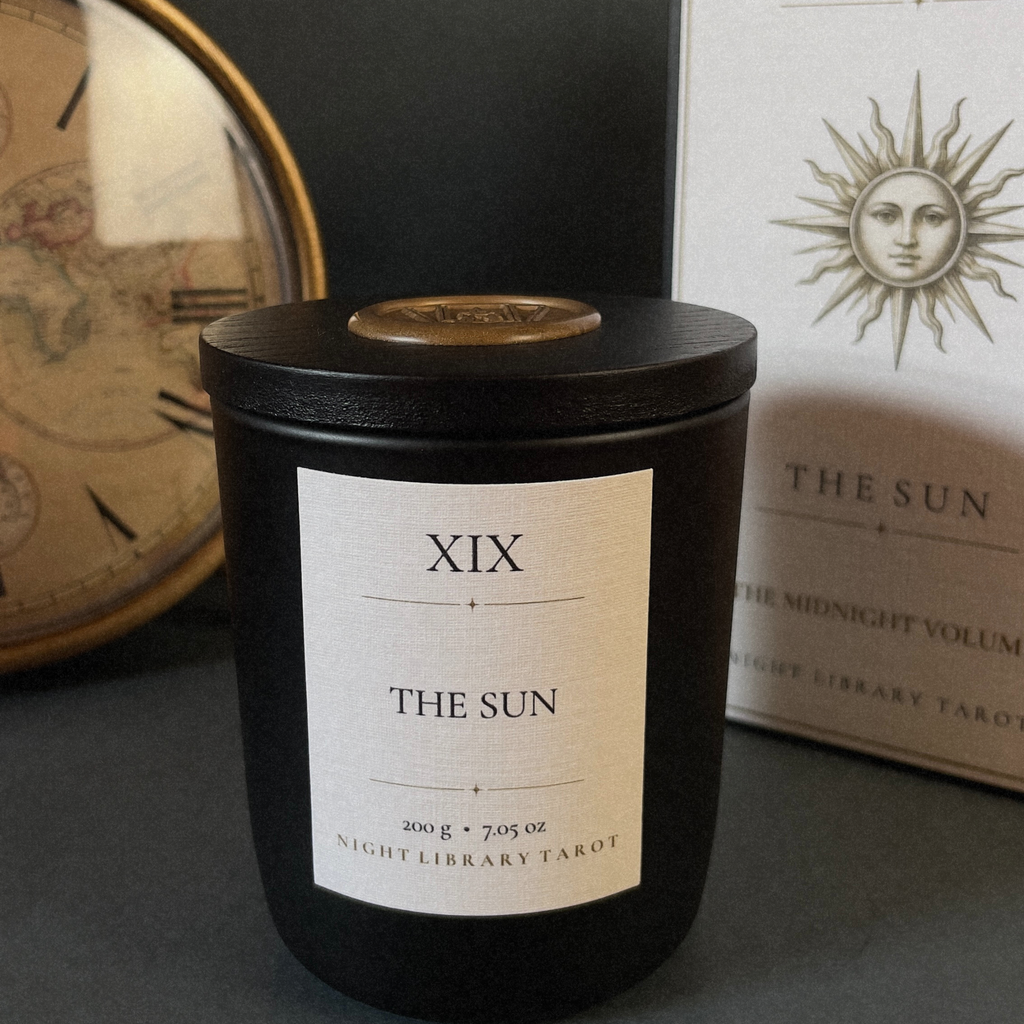 A black glass jar candle labeled 'XIX THE SUN' sits on a dark surface, with part of its white box visible behind it and a vintage-style clock in the background.
