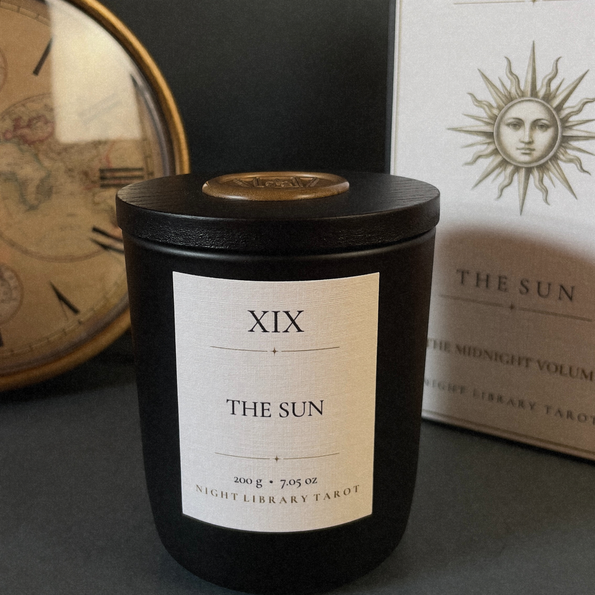 A black glass jar candle labeled 'XIX THE SUN' sits on a dark surface, with part of its white box visible behind it and a vintage-style clock in the background.