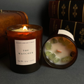 An amber glass candle jar labeled 'Night Library Tarot - The Watcher' with a lit flame, shown beside its open lid revealing green stone inclusions in white wax, with vintage books in the background.