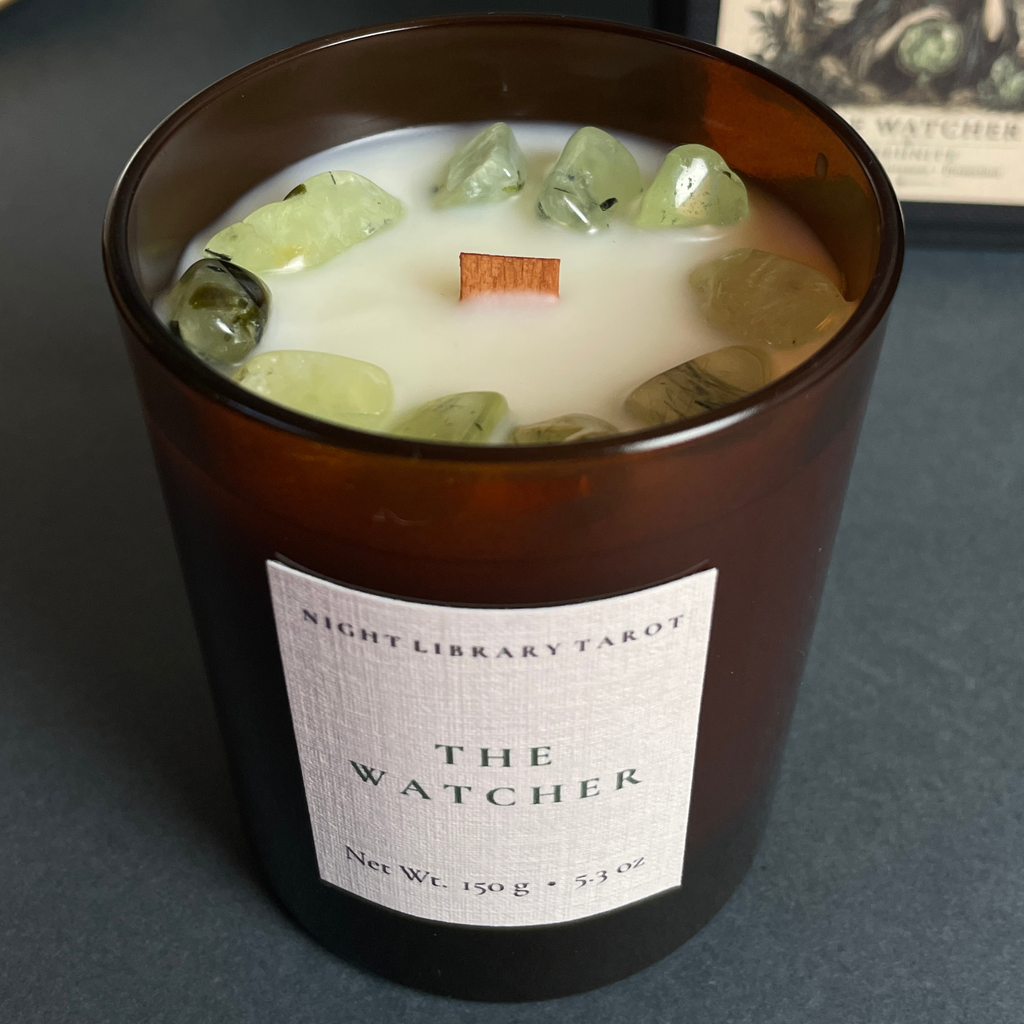 An amber glass jar candle with white wax and green crystal stones on top, featuring a white label that reads 'Night Library Tarot The Watcher'.